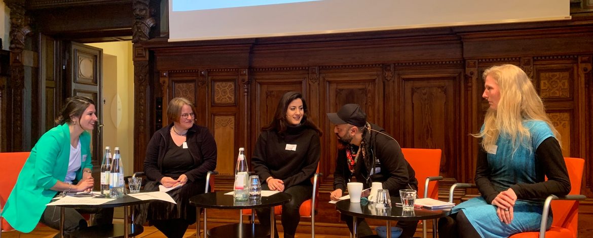 Auf diesem Foto befinden sich die vier Paneldiskutant*innen und die Moderation aus dem Podium